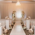 Elegant indoor wedding ceremony with white aisle runner, crystal chandelier, ivory draping, tall floral columns, and candlelit white chiavari chairs