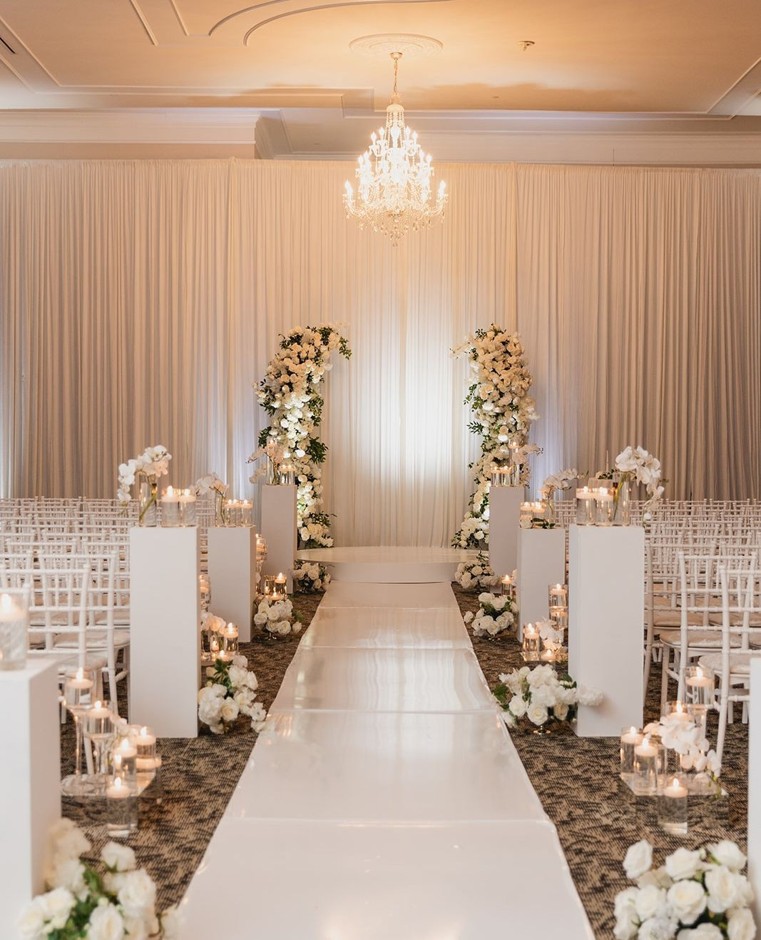 Elegant indoor wedding ceremony with white aisle runner, crystal chandelier, ivory draping, tall floral columns, and candlelit white chiavari chairs