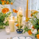 Wedding table centerpiece featuring orange marigolds, white flowers, cylinder candles, and blue-patterned bowl with blue-rimmed plates