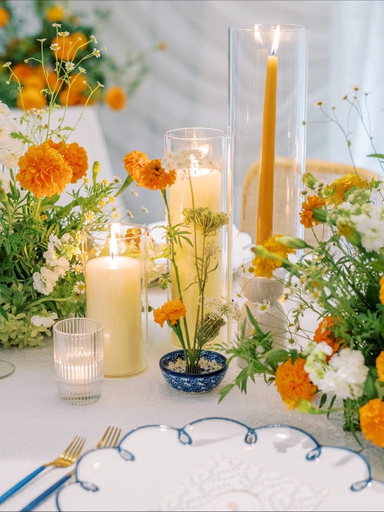 Wedding table centerpiece featuring orange marigolds, white flowers, cylinder candles, and blue-patterned bowl with blue-rimmed plates