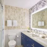 Modern bathroom with navy vanity, backlit mirror, travertine tile walls, and decorative wallpaper accent