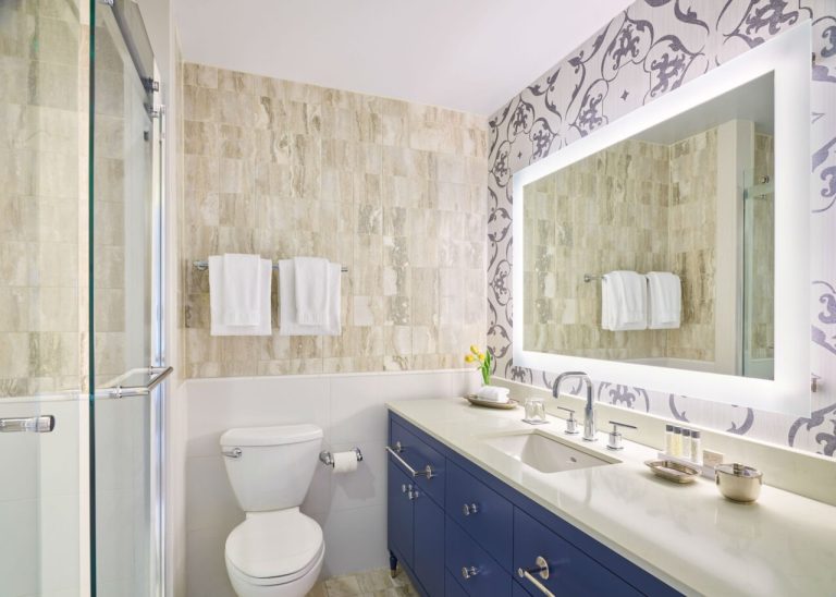 Modern bathroom with navy vanity, backlit mirror, travertine tile walls, and decorative wallpaper accent