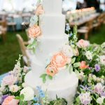 Five-tier white wedding cake adorned with cascading peach and blush roses displayed at an outdoor tented reception surrounded by fresh flowers