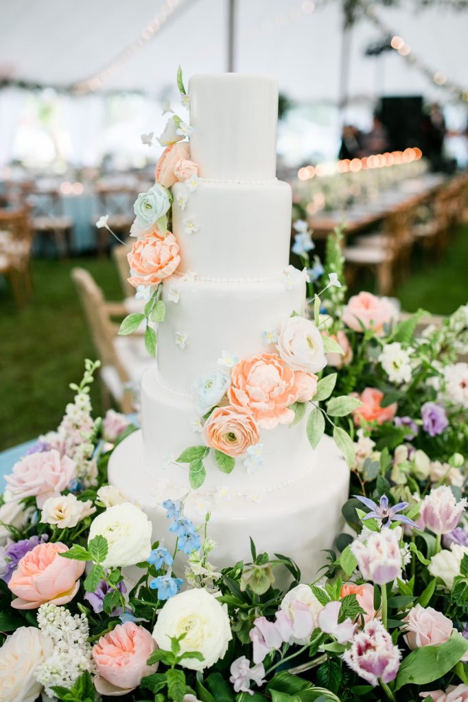 Five-tier white wedding cake adorned with cascading peach and blush roses displayed at an outdoor tented reception surrounded by fresh flowers