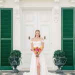 Bride in white gown with colorful bouquet poses on checkered patio in front of colonial-style entrance
