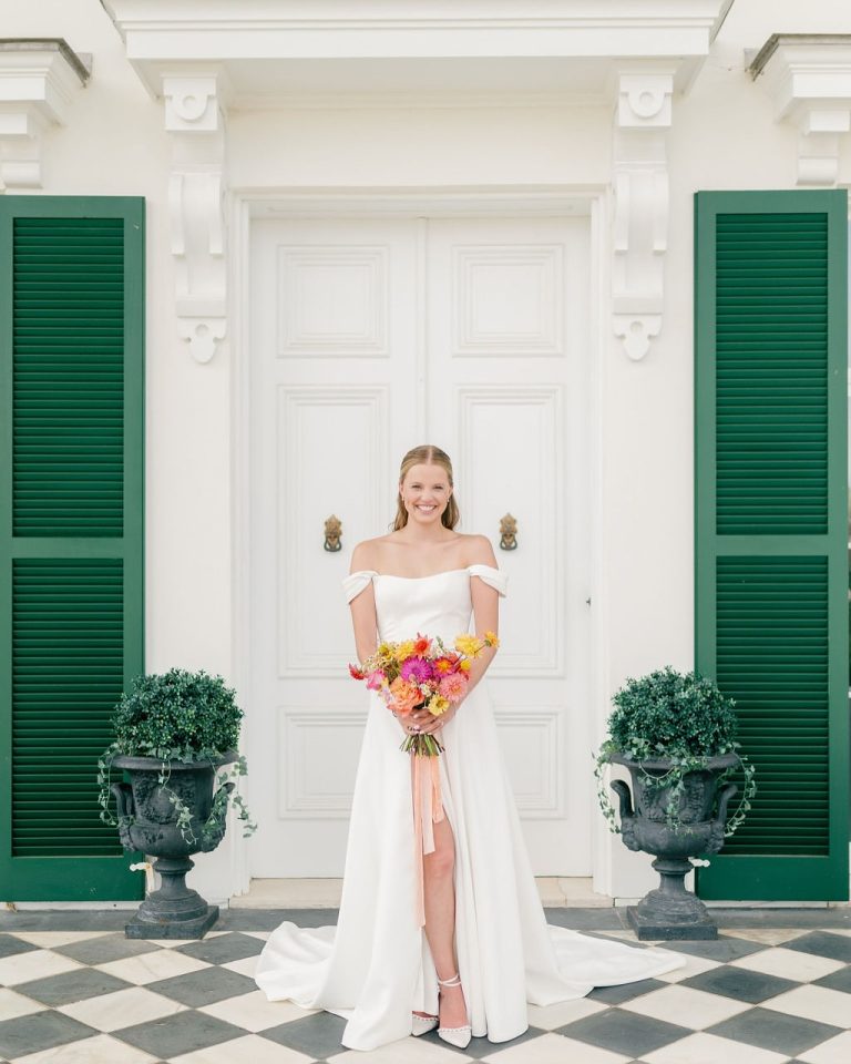 Bride in white gown with colorful bouquet poses on checkered patio in front of colonial-style entrance