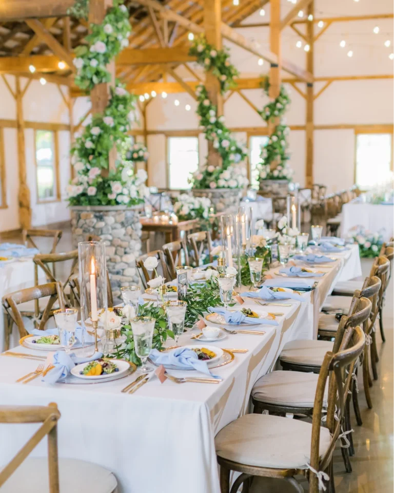 Rustic barn wedding reception tables with greenery garlands, tall candles, and blue napkins