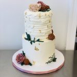 Two-tier white wedding cake with textured buttercream, gold leaf accents, and burgundy roses on pink stand