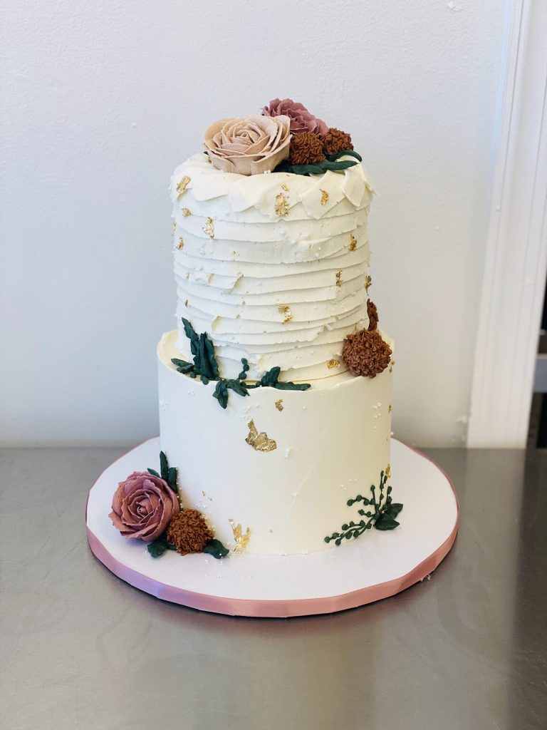 Two-tier white wedding cake with textured buttercream, gold leaf accents, and burgundy roses on pink stand