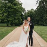 Bride and groom stand together on tree-lined pathway in formal wedding attire