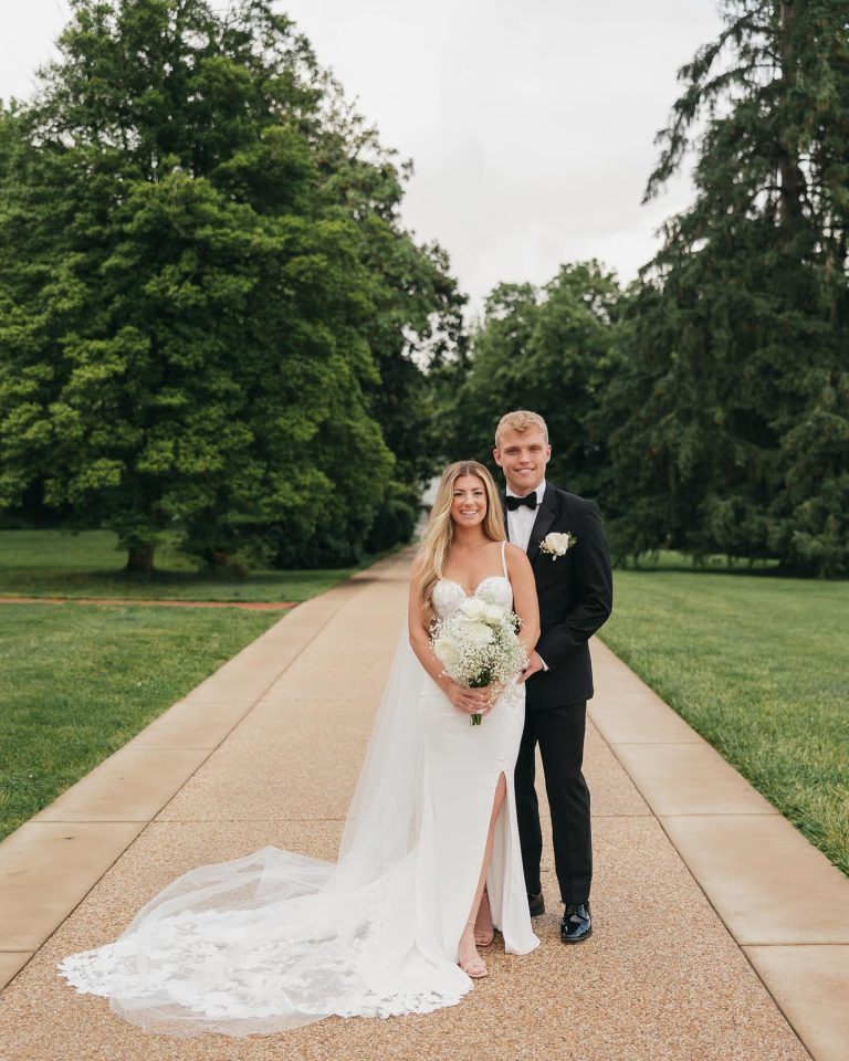 Bride and groom stand together on tree-lined pathway in formal wedding attire