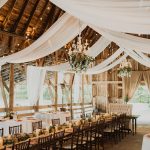 Elegant barn wedding venue interior featuring exposed wood beams, flowing ceiling drapes, and chandelier lighting