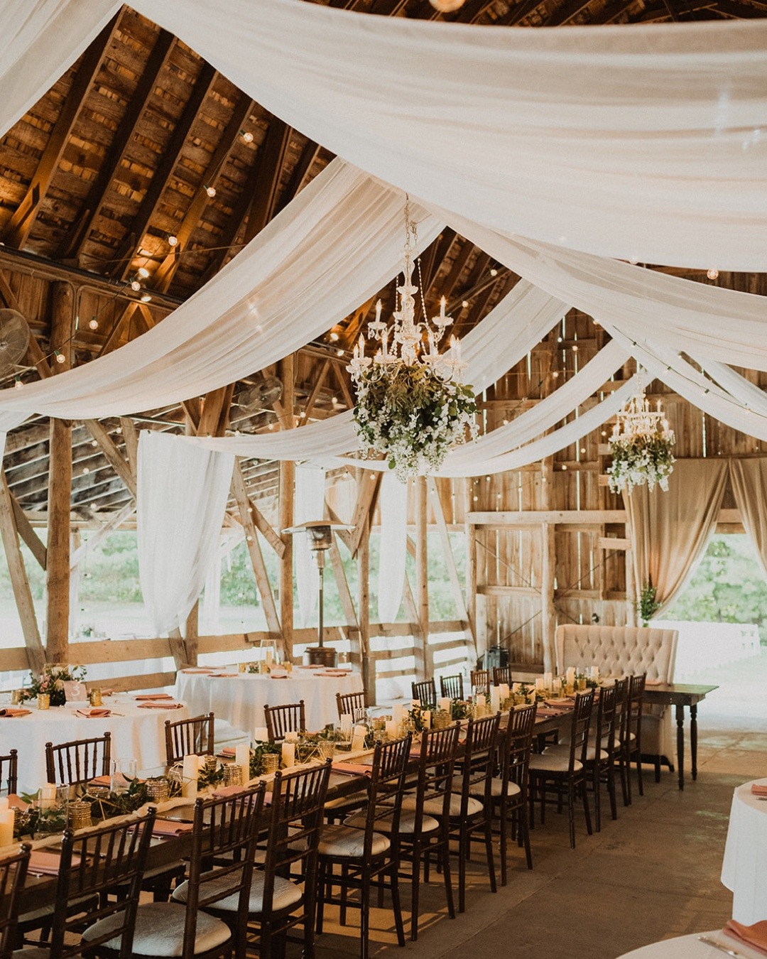 Elegant barn wedding venue interior featuring exposed wood beams, flowing ceiling drapes, and chandelier lighting