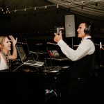 Wedding DJ in headphones filming bride dancing at outdoor reception with string lights