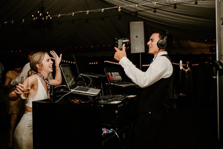 Wedding DJ in headphones filming bride dancing at outdoor reception with string lights
