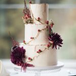 Three-tier white wedding cake with gold-trimmed edges, adorned with burgundy dahlias, red berries, and trailing amaranthus on a glass cake stand