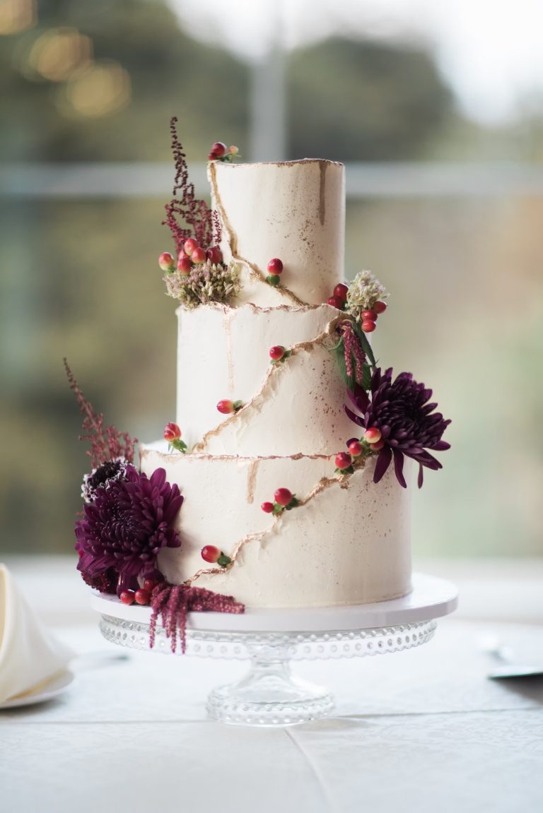 Three-tier white wedding cake with gold-trimmed edges, adorned with burgundy dahlias, red berries, and trailing amaranthus on a glass cake stand