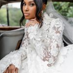 Bride in white floral lace gown and veil poses elegantly in vintage car on wedding day