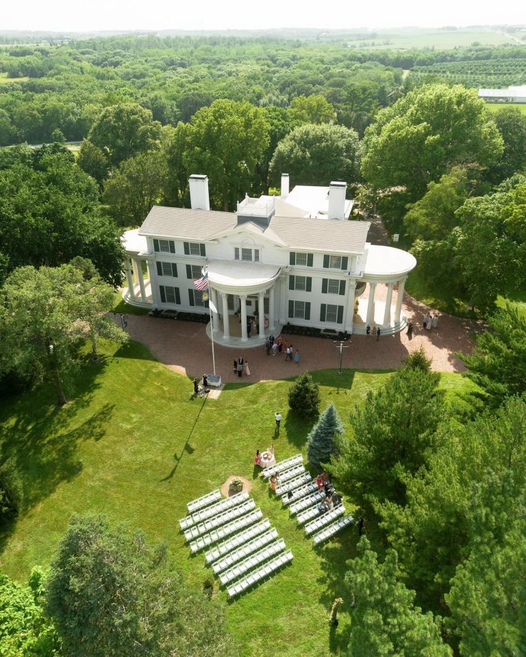 Aerial view of white columned mansion wedding venue with ceremony seating on manicured lawn surrounded by trees
