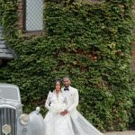 Bride and groom pose beside vintage white car in front of ivy-covered stone building with cathedral veil flowing