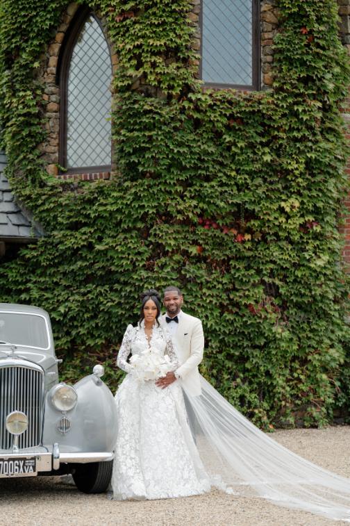 Bride and groom pose beside vintage white car in front of ivy-covered stone building with cathedral veil flowing