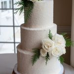 Three-tier white frosted wedding cake with sugar coating, decorated with ivory and blush roses, eucalyptus, and fern on wood slice stand