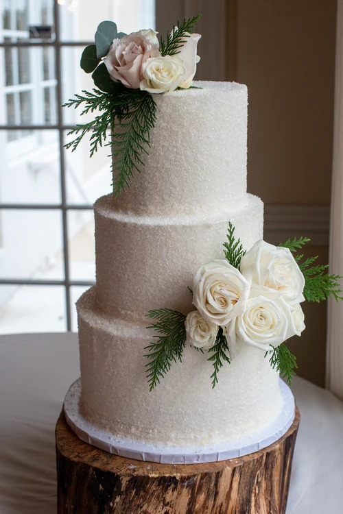 Three-tier white frosted wedding cake with sugar coating, decorated with ivory and blush roses, eucalyptus, and fern on wood slice stand