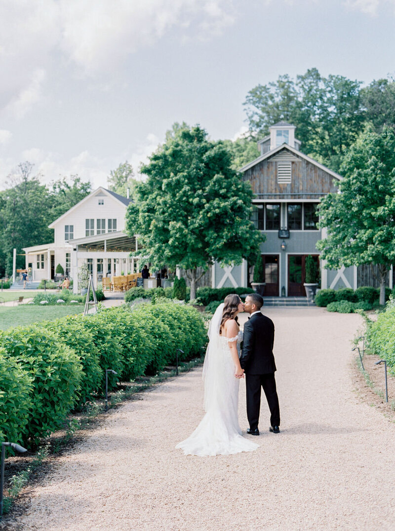 Bride and groom kissing on gravel path at rustic Northern Virginia farm wedding venue with barn buildings