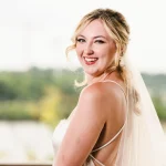 Bride with natural makeup and loose updo smiling over shoulder by waterfront