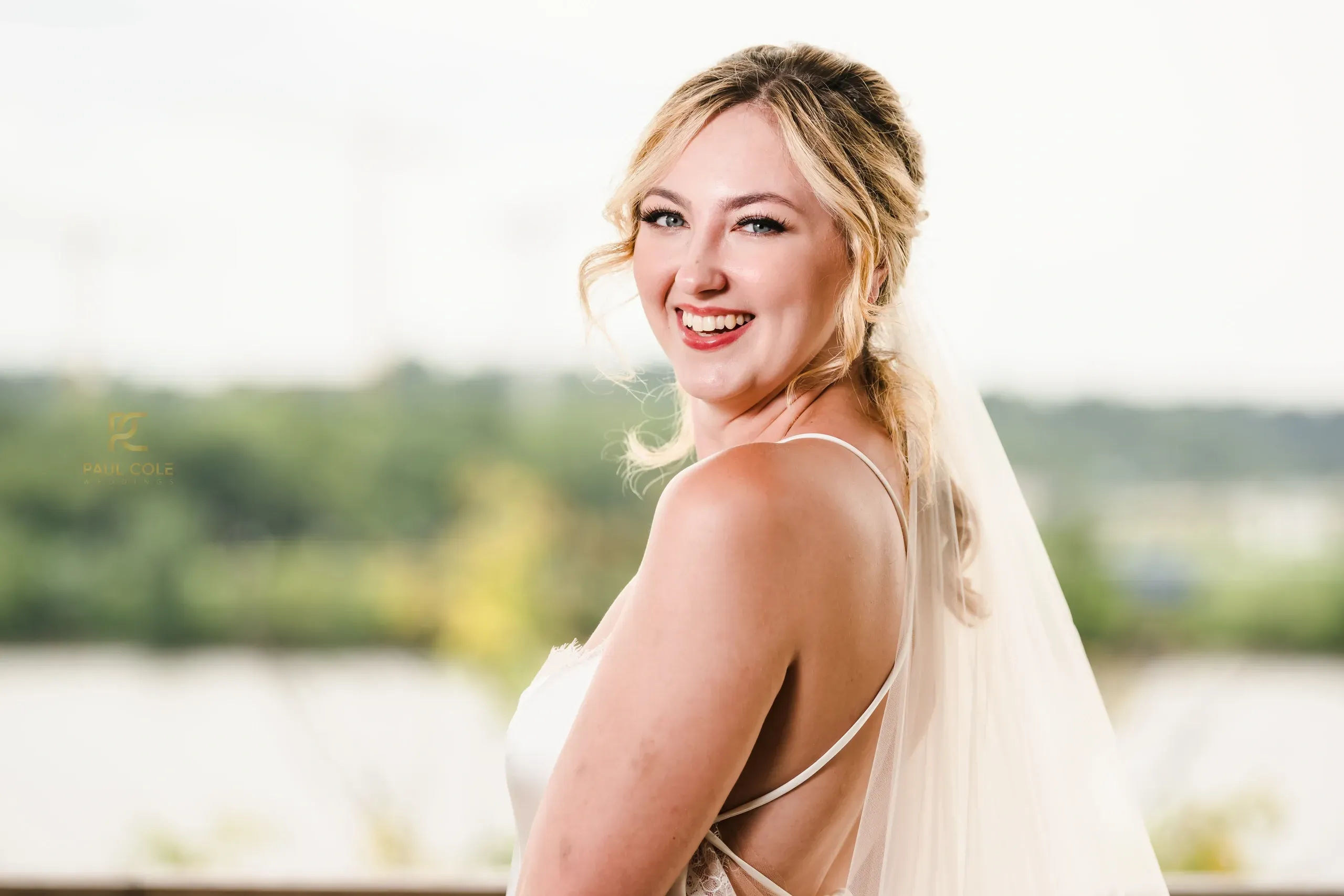 Bride with natural makeup and loose updo smiling over shoulder by waterfront