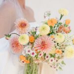Bride wearing veil holds pastel bouquet with pink dahlias, yellow carnations, orange poppies, and white blooms