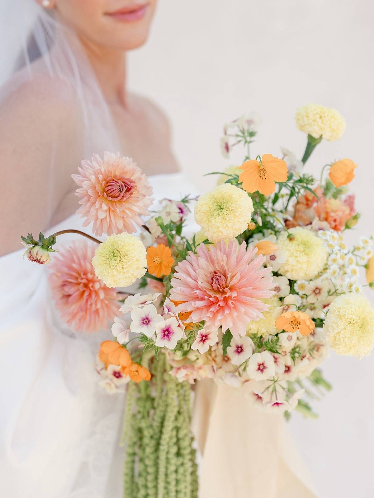 Bride wearing veil holds pastel bouquet with pink dahlias, yellow carnations, orange poppies, and white blooms