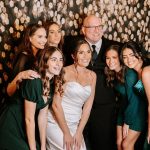 Bride in white gown with wedding party in emerald green dresses posing at photo booth with gold sequin backdrop