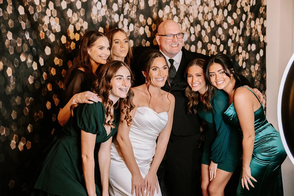 Bride in white gown with wedding party in emerald green dresses posing at photo booth with gold sequin backdrop
