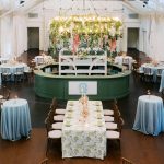 Elegant event space with blue tablecloths, circular bar with floral chandelier, and long head table under white tent