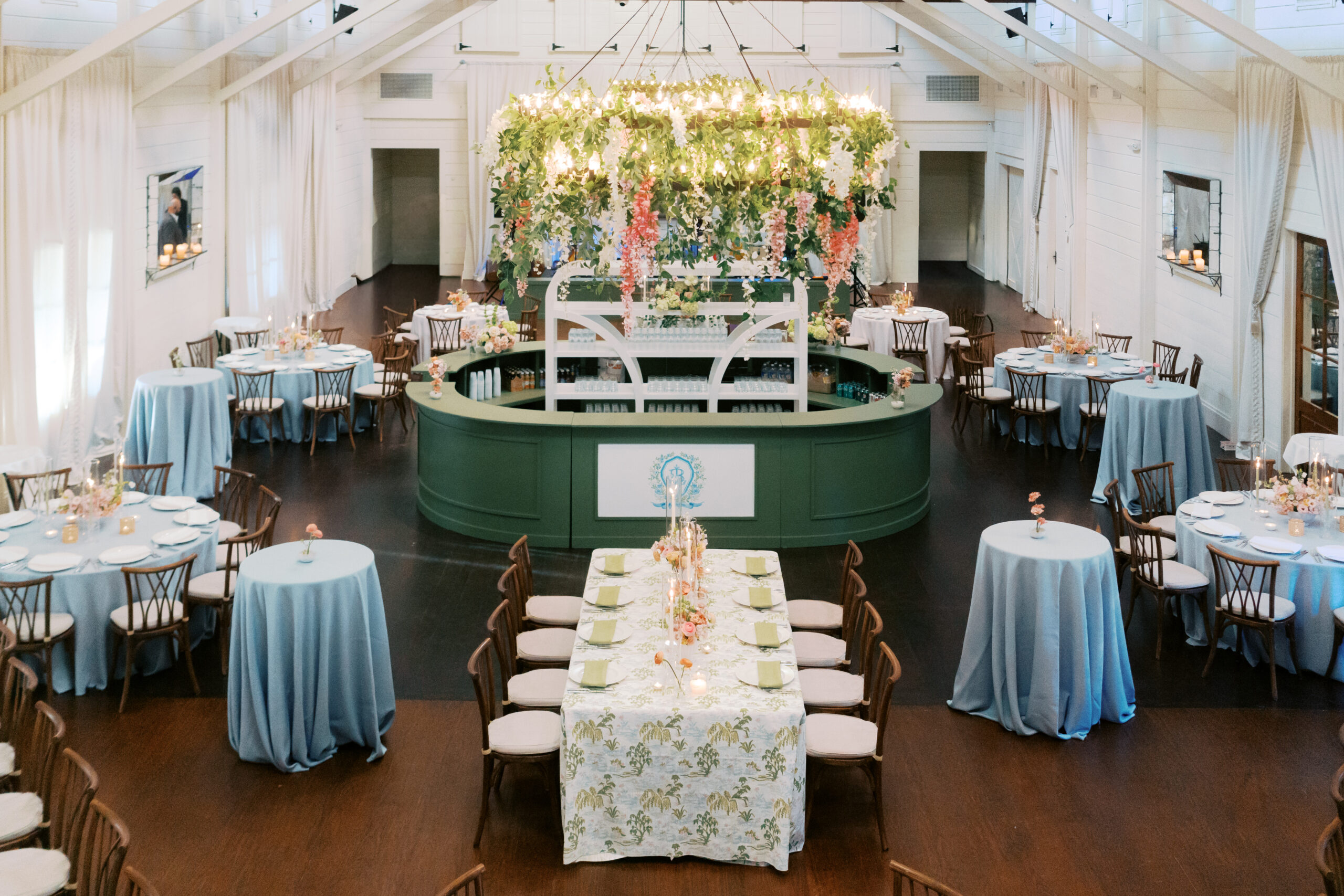 Elegant event space with blue tablecloths, circular bar with floral chandelier, and long head table under white tent