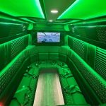 Spacious party bus interior featuring green LED lighting, quilted leather bench seating, and flat-screen TV