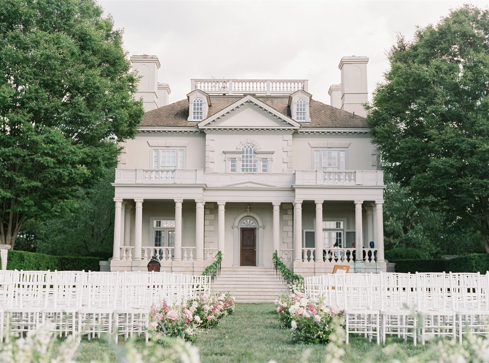 White neoclassical mansion with columned portico and outdoor wedding ceremony setup with white chairs on manicured lawn