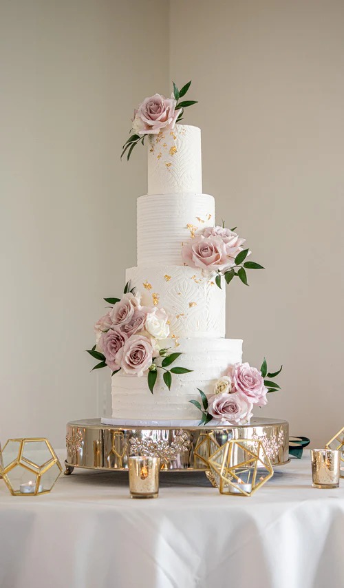 Four-tier white wedding cake with horizontal texture, gold leaf accents, and dusty pink roses on a mirrored stand with gold geometric candle holders