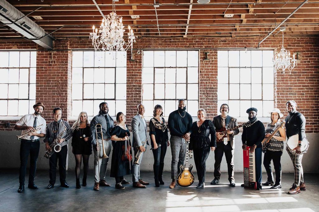 Diverse wedding band posing with instruments in brick loft venue with crystal chandeliers and industrial windows