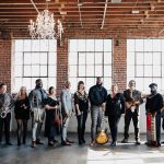 Diverse wedding band posing with instruments in brick loft venue with crystal chandeliers and industrial windows
