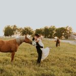 Newlyweds kissing beside horse at golden hour on ranch property with white fencing and barn