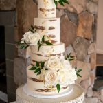 Four-tier semi-naked wedding cake with exposed layers, white roses, and greenery on ornate white pedestal