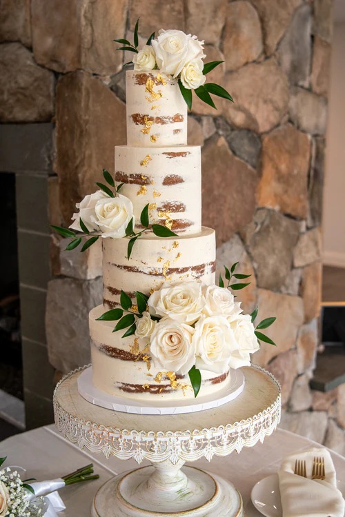 Four-tier semi-naked wedding cake with exposed layers, white roses, and greenery on ornate white pedestal