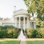 Newlyweds embracing on mansion lawn with stately white columns and gardens behind them