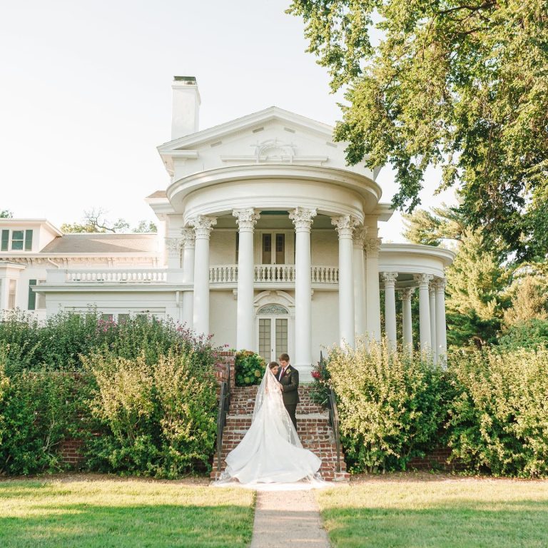 Newlyweds embracing on mansion lawn with stately white columns and gardens behind them