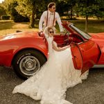 Couple in vintage wedding attire with classic red Corvette beneath golden-hour tree canopy