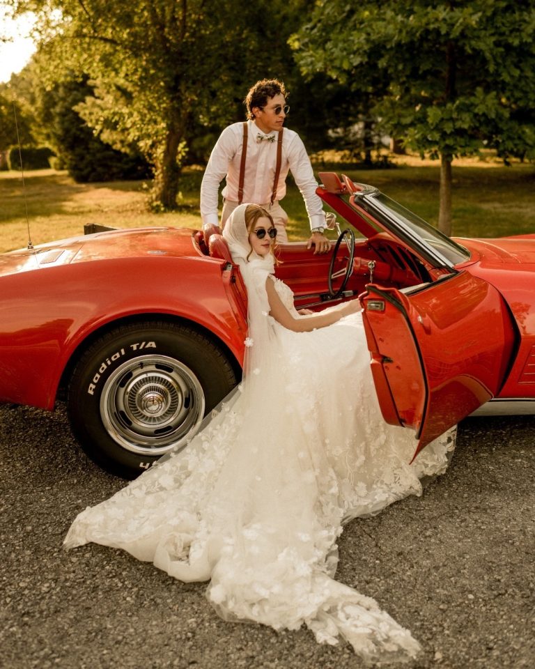 Couple in vintage wedding attire with classic red Corvette beneath golden-hour tree canopy