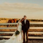 Bride and groom pose by white fence at outdoor Nebraska wedding venue with horses and pasture backdrop
