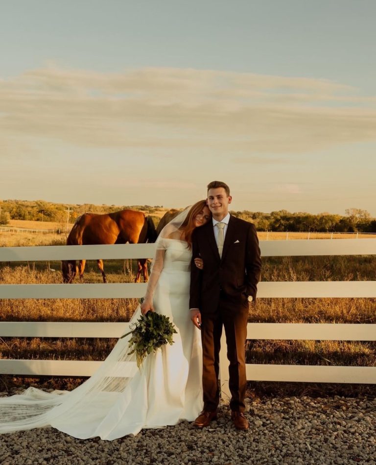 Bride and groom pose by white fence at outdoor Nebraska wedding venue with horses and pasture backdrop